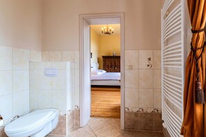 Un bany amb vàter, dutxa i terra de fusta que condueix a una habitació amb un llit i una tauleta de nit.