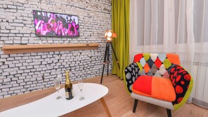 Ein modernes Wohnzimmer mit Backsteinwand, einem Wand-Fernseher, einem Tisch mit Flasche und Gläsern, einem farbenfrohen Sessel und einer Stehlampe.