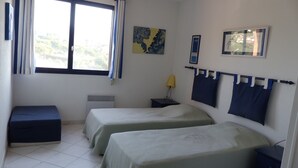Ein Zimmer mit zwei Betten, einem Fenster mit Vorhängen, einem Heizkörper, einem kleinen Tisch mit Lampe und einer Wand mit gerahmten Bildern.