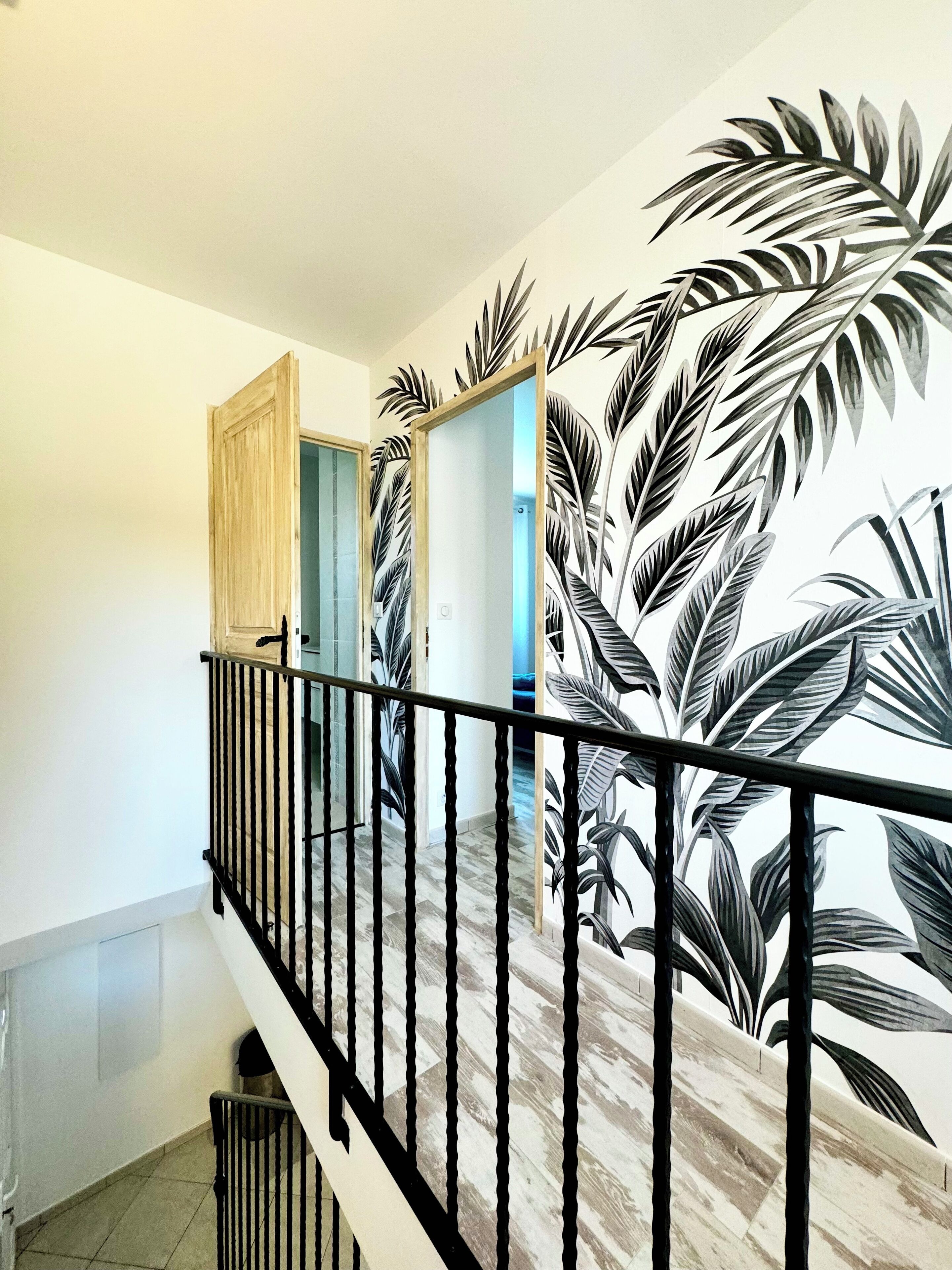 Un escalier avec une rampe noire, une porte en bois et un mur orné d’un motif de feuilles tropicales.