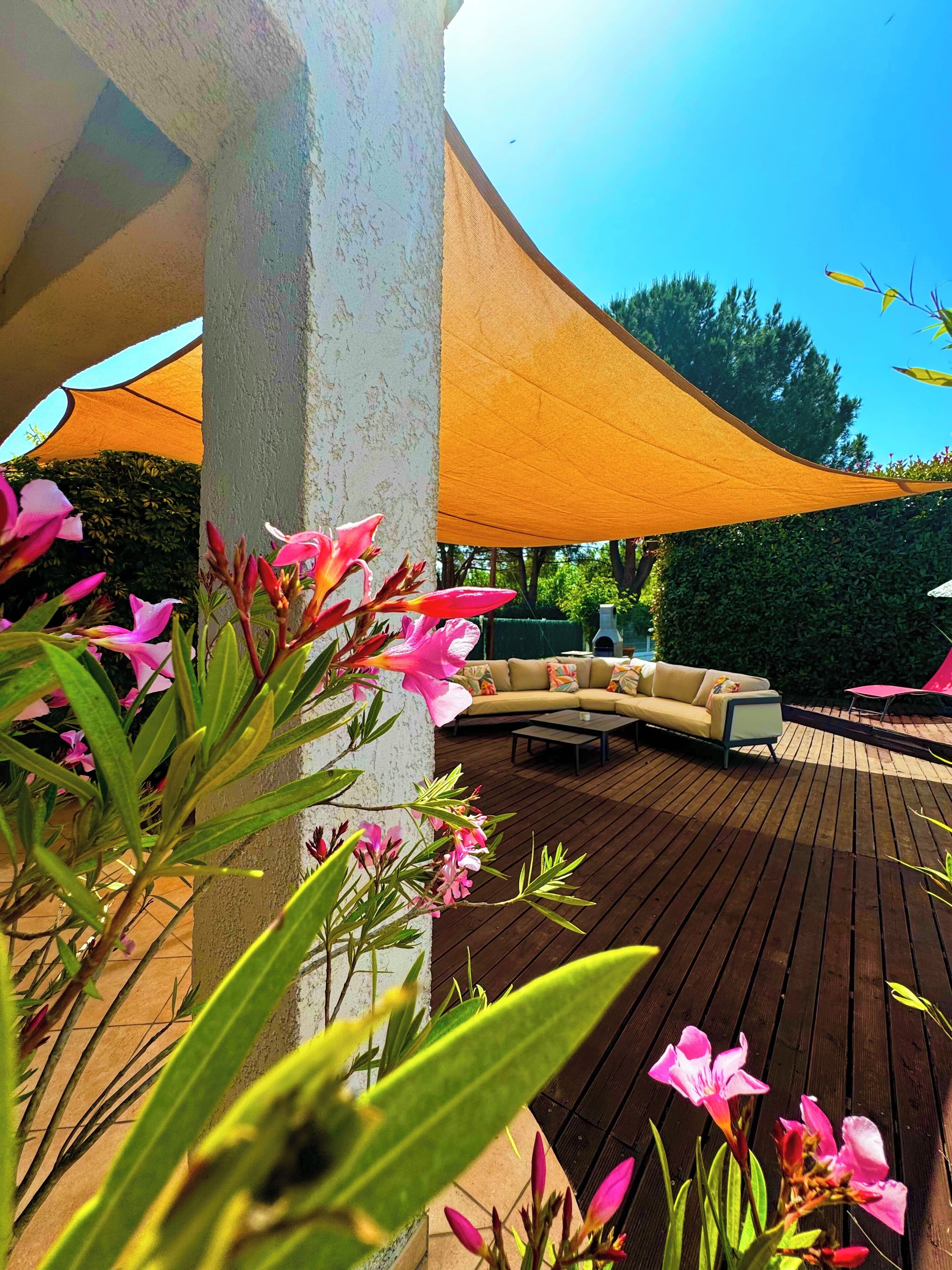 Une terrasse en bois avec un canapé beige et une table basse, entourée de fleurs roses et protégée par une grande voile d’ombrage.