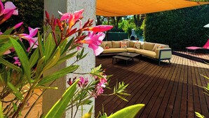 A wooden deck with a beige sofa and a coffee table, surrounded by pink flowers and a large shade sail. - Campo Vecchio DRACENA (Ghisonaccia)