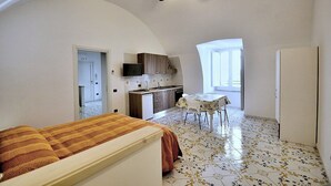 Een compacte kamer met een bed, een kitchenette, een eettafel en een plafondventilator.