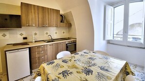Una cocina compacta con armarios de madera, un lavamanos y un refrigerador pequeño. Hay una zona de comedor con una mesa cubierta por un mantel estampado con hojas.
