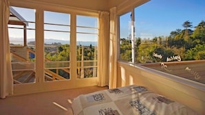 Une chambre avec de grandes fenêtres, un lit recouvert d’une courtepointe à motifs et une vue sur un quartier résidentiel.