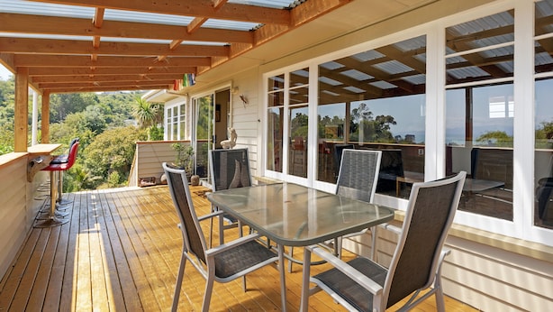 Une terrasse extérieure couverte, dotée d’une table et de chaises en verre, d’un plancher en bois et de grandes fenêtres offrant une vue sur la verdure environnante.