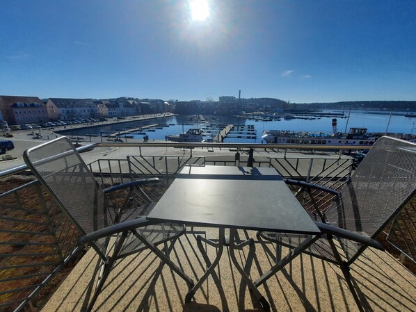 A view from a high vantage point overlooking a marina with boats docked, a clear sky, and a table with chairs in the foreground.