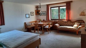 Una habitación con una cama, una mesa de comedor con sillas, un sofá, una ventana con cortinas y un radiador.