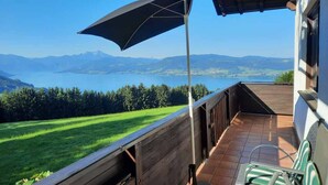 Un balcón con vistas a un lago y montañas, baranda de madera y una silla.