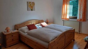 Ein hölzernes Bett mit zwei Kissen, ein Nachttisch mit Lampe und ein Fenster mit Vorhängen.