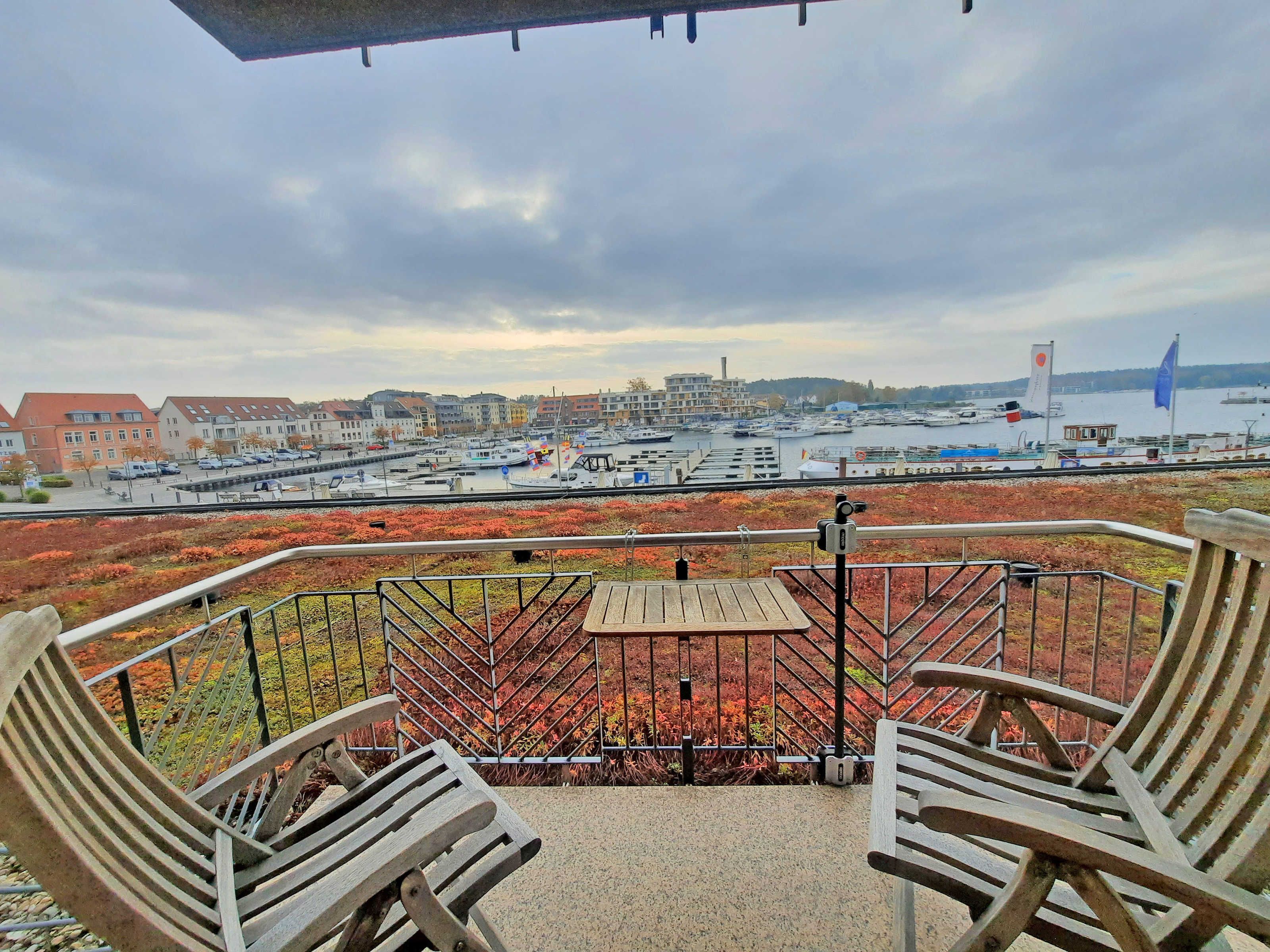 A balcony with two wooden chairs, a table, and a view of a marina with boats and buildings in the background.
