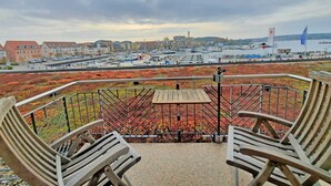 Een balkon met twee houten stoelen, een tafel en uitzicht op een jachthaven met boten en gebouwen op de achtergrond.