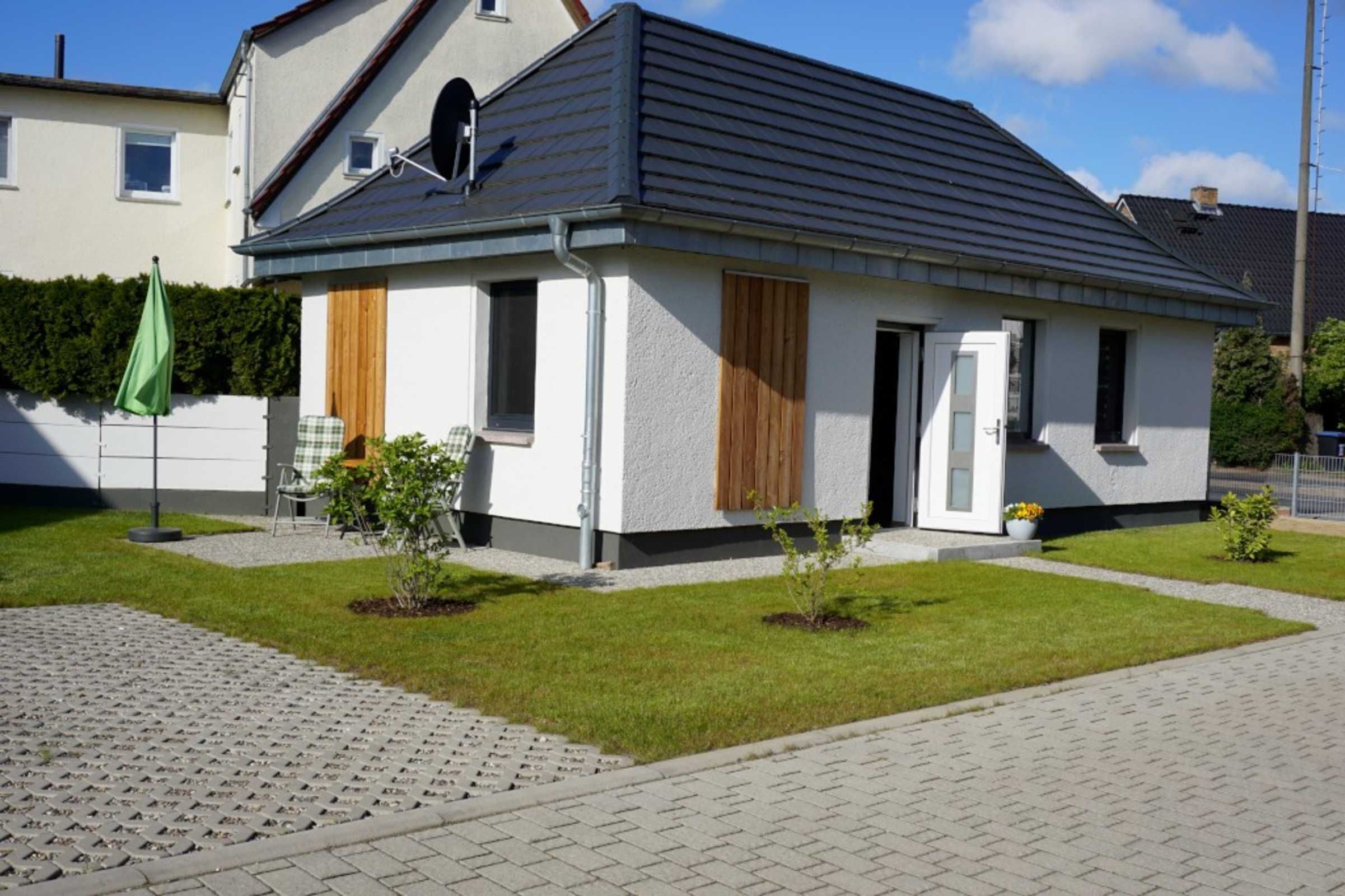 Ein einstöckiges Haus mit grauem Dach, weißen Außenwänden und einem kleinen Vorgarten mit gepflasterter Auffahrt.