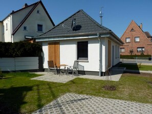 Une petite maison de plain-pied, avec un toit en pente, une porte en bois et un petit patio comprenant une table et des chaises.