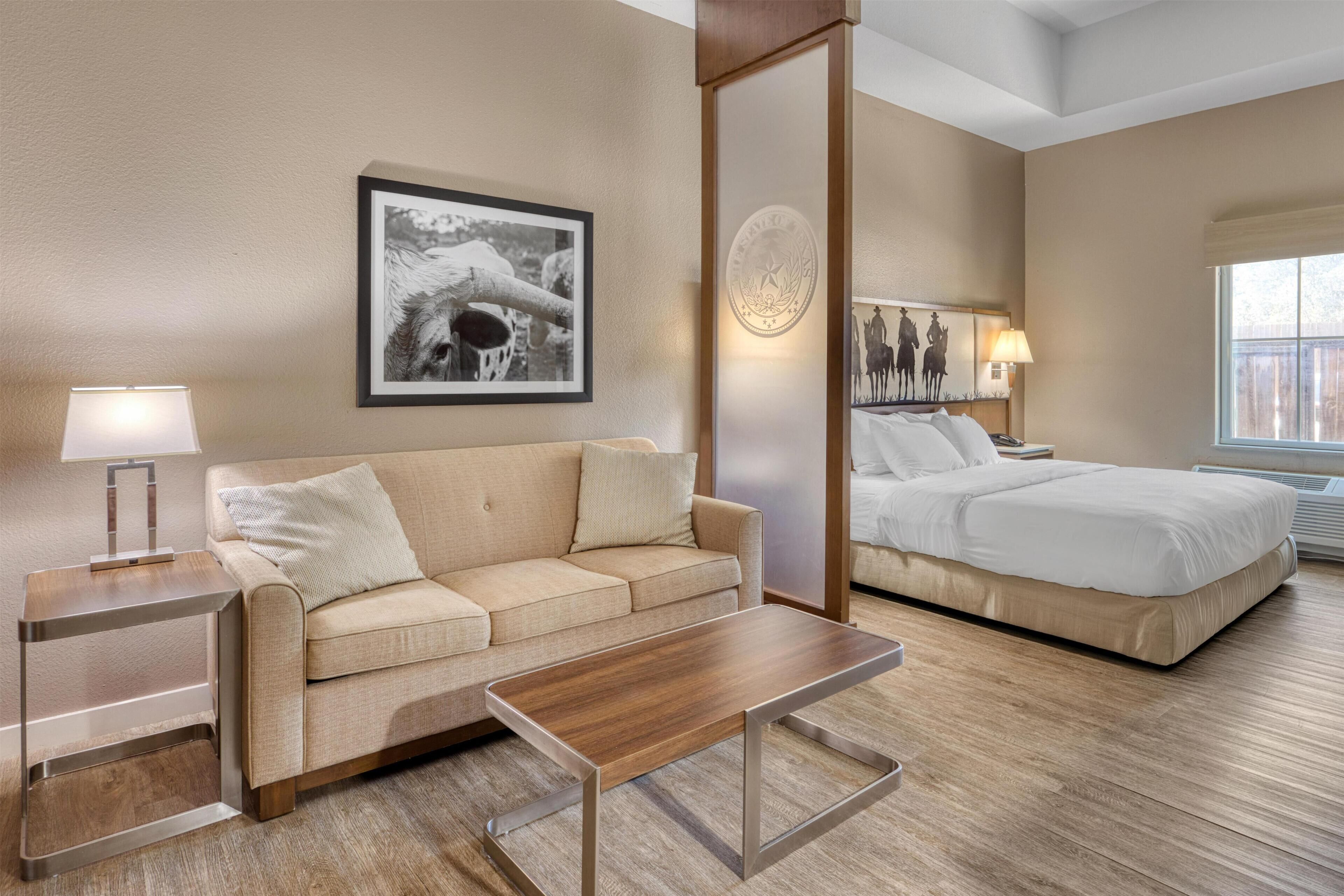 A hotel room with a sofa, a wooden coffee table, a bed, a nightstand, a lamp, and a framed picture on the wall.