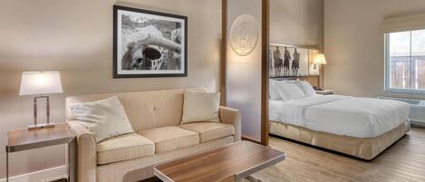 A hotel room with a sofa, a wooden coffee table, a bed, a nightstand, a lamp, and a framed picture on the wall.