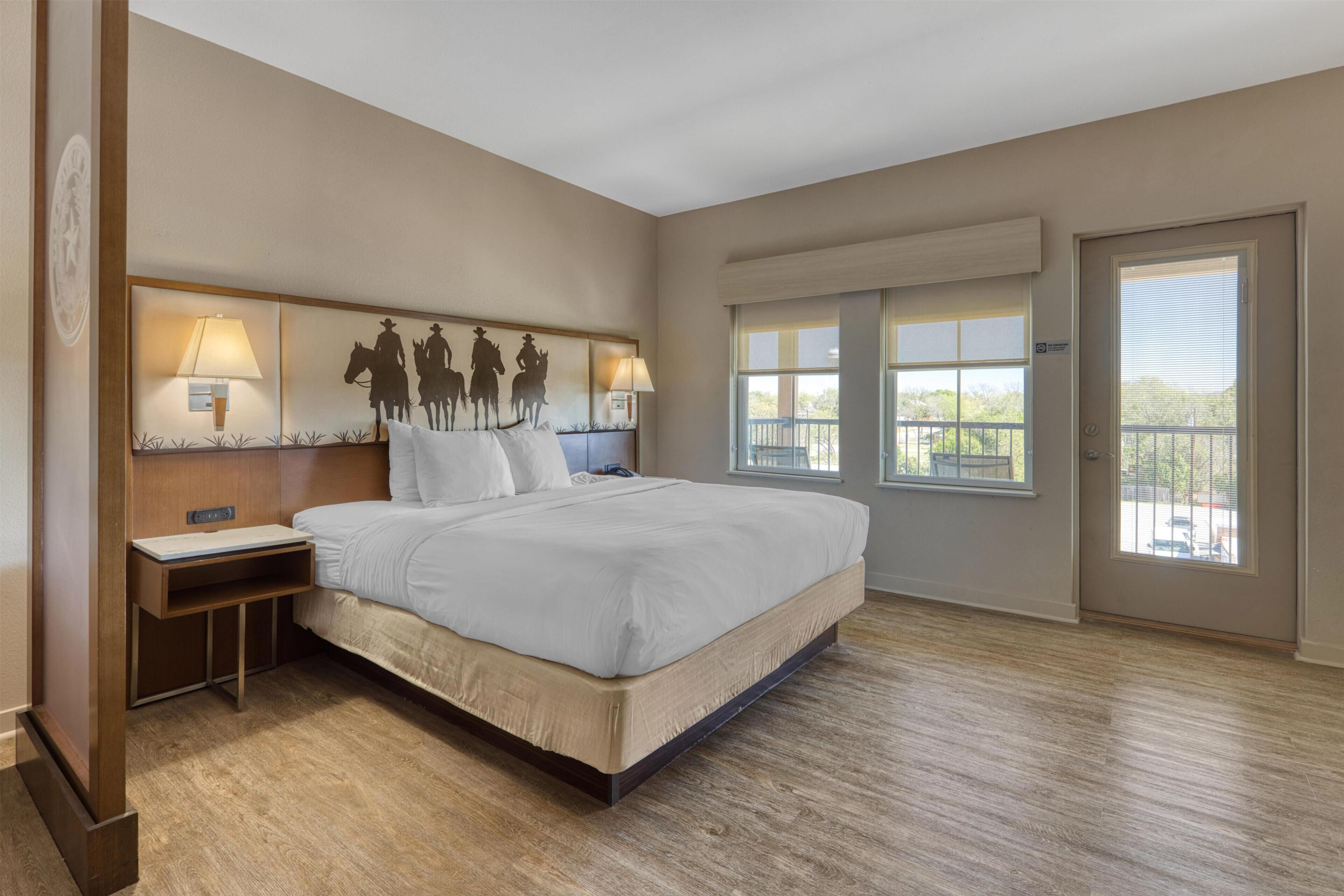 A hotel room with a large bed, bedside lamps, a nightstand, a headboard with a silhouette artwork, a window with a view, and a door with a window.