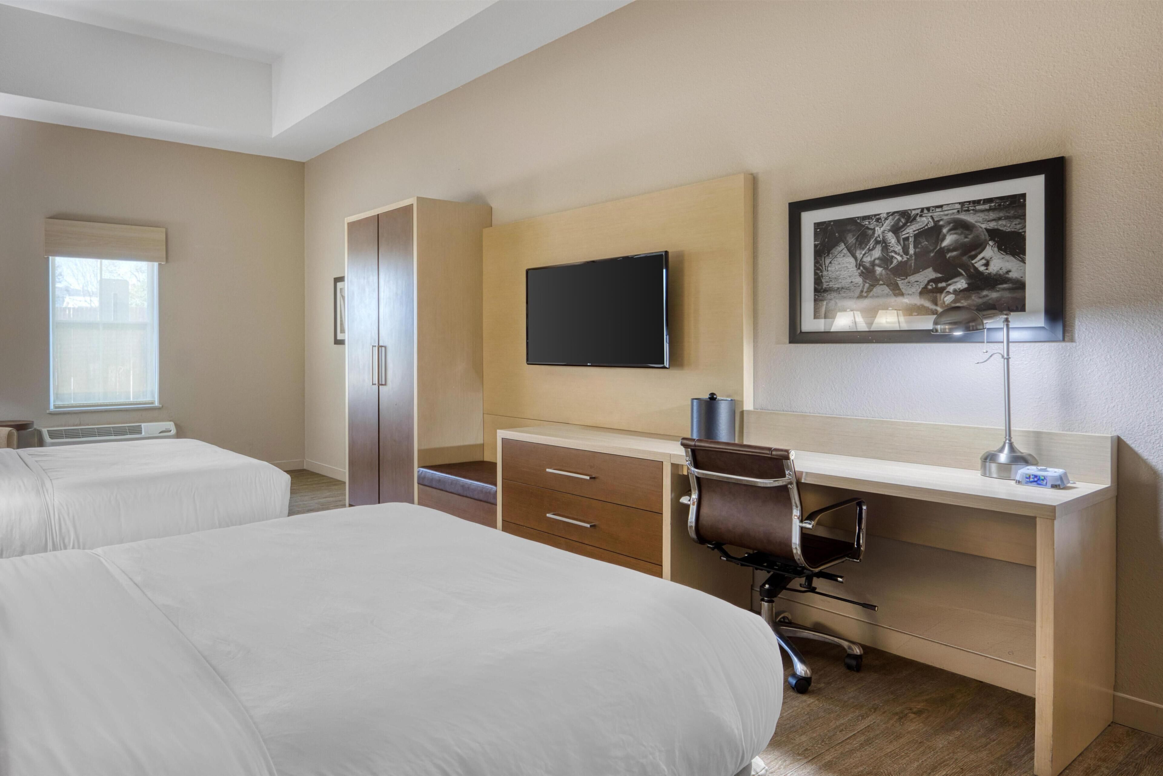 A hotel room with two beds, a desk with a chair, a flat-screen TV, a framed picture on the wall, and a window with blinds.