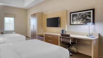A hotel room with two beds, a desk with a chair, a flat-screen TV, a framed picture on the wall, and a window with blinds.