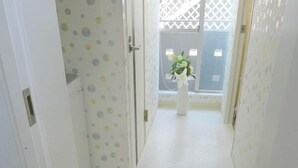 A narrow hallway with a white door on the left, a white wall with a polka dot pattern, and a white bench leading to a window with a white vase and green plants. - Pacio Nijo (Kyoto)