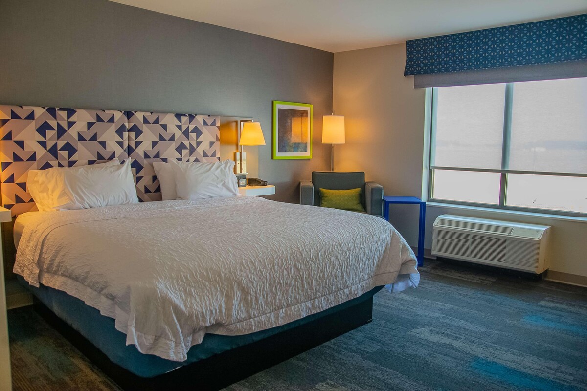 A hotel room with a large bed, two bedside lamps, a chair, a window with blinds, and a wall-mounted air conditioner.