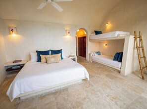 Une chambre avec un grand lit, un lit superposé, une table de chevet, une lampe et un ventilateur de plafond.