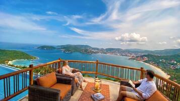 Un balcon agrémenté de meubles en osier et d’une bougie, offrant une vue sur la côte, avec une piscine et une plage.
