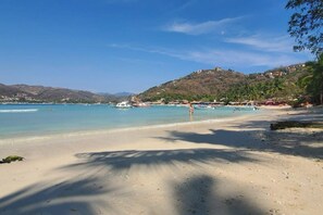 Une plage à l’eau claire, avec des bateaux et une chaîne de collines en arrière-plan.
