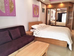 A hotel room with a bed, a wooden nightstand, a purple sofa, and a small table. - Spacious 3rd Floor Studio at Baan Suan Lalana (Pattaya)