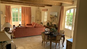 En hyggelig stue med sofa, spisebord, stole, træbjælker i loftet og store vinduer med gardiner.