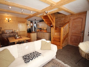 Een gezellig interieur van een blokhut met een houten eettafel, een bank met decoratieve kussens, een trap en een keukenhoek.