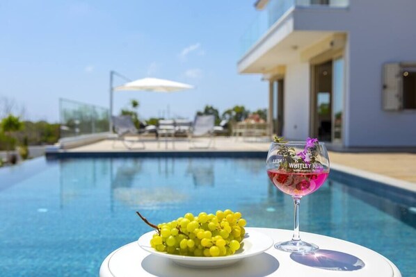 A poolside setting with a bowl of yellow grapes and a glass of red wine on a white table.