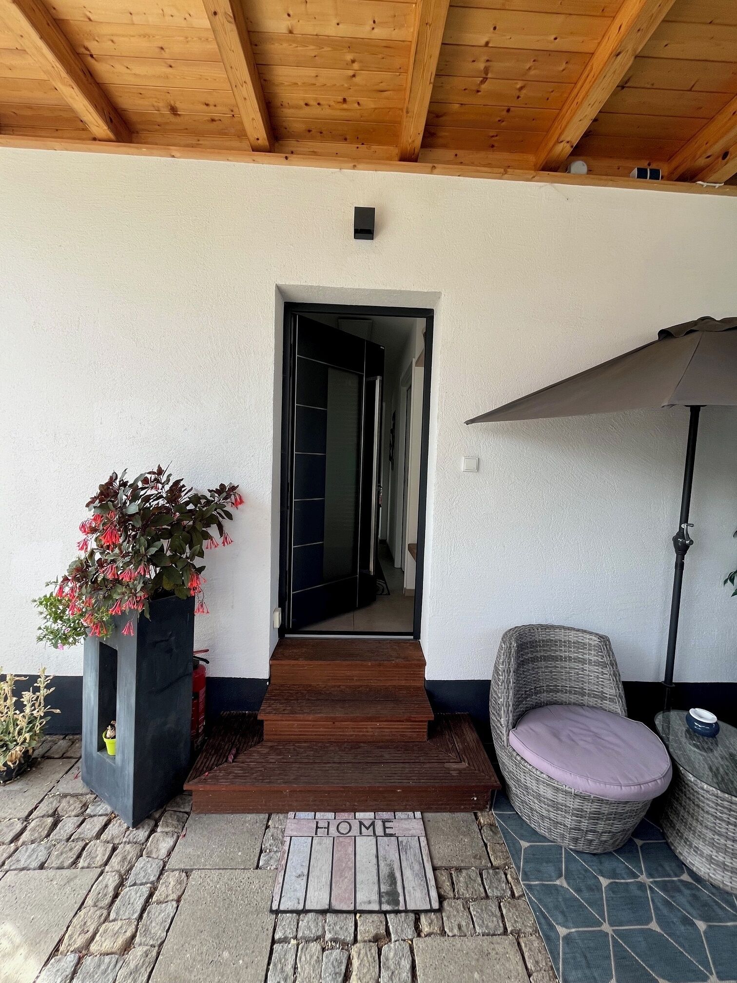 Une entrée abritée avec un plafond en bois, une plante en pot, une chaise en osier et un paillasson sur lequel est écrit le mot « MAISON ».