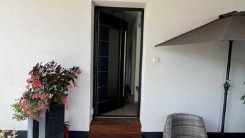 Une entrée abritée avec un plafond en bois, une plante en pot, une chaise en osier et un paillasson sur lequel est écrit le mot « MAISON ».