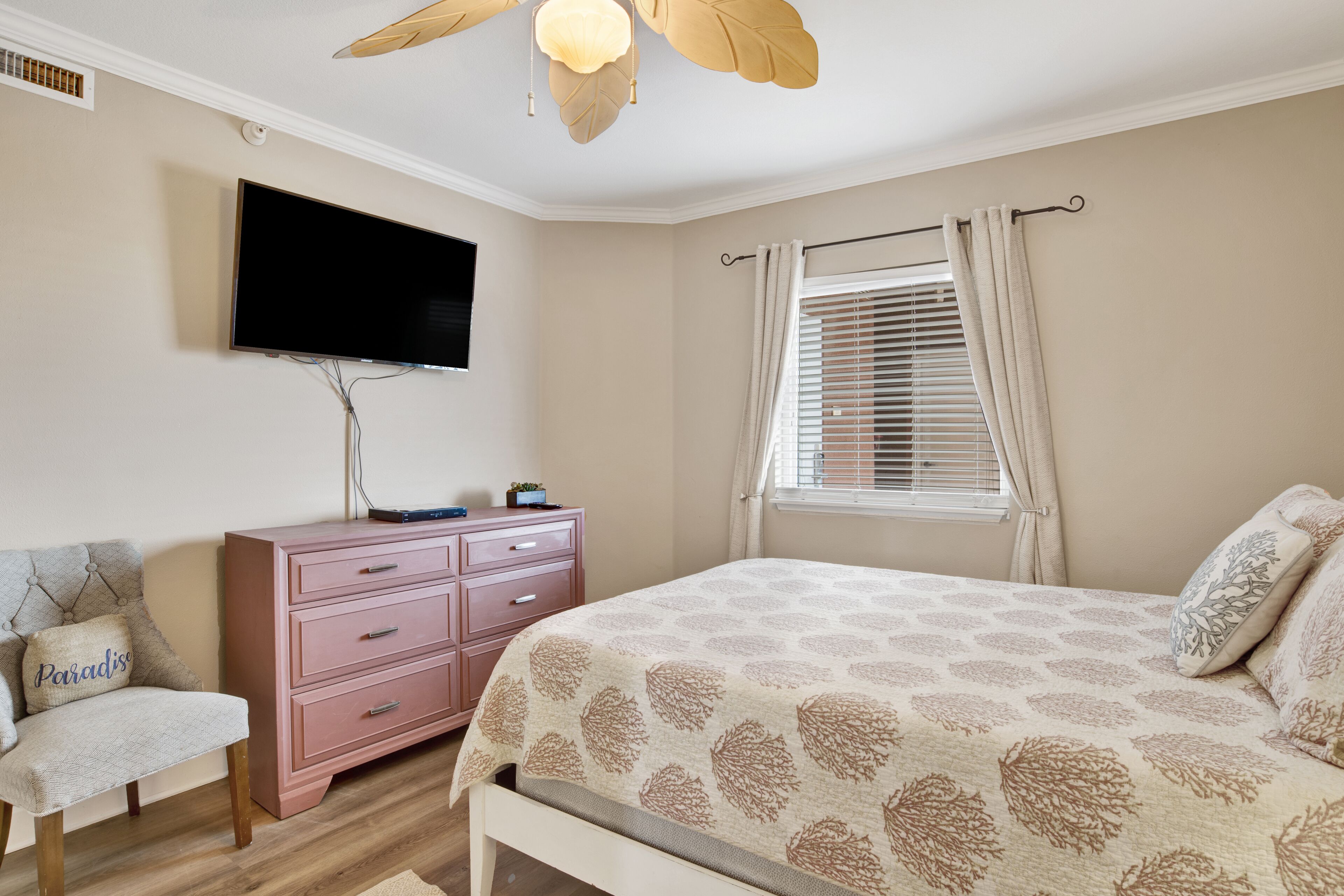 Un dormitorio con cama, cómoda, silla, televisor y ventana con cortinas.