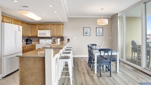 Eine moderne Küche mit Holzschränken, einem weißen Kühlschrank, einer Mikrowelle und einer Kaffeemaschine. Es gibt eine Essecke mit Tisch und Stühlen, und ein großes Fenster mit Blick nach draußen.