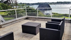 An outdoor patio with wicker furniture, a white railing, and a view of a lake and buildings in the background. - Grande Maison Avec vue Imprenable sur L'aber-wrac'h (Plouguerneau)