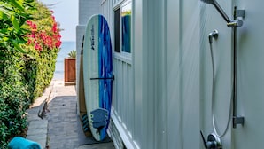 Ein schmaler Pfad führt zu einem Gebäude mit einer Duschanlage, an der eine Surfboard angelehnt ist.