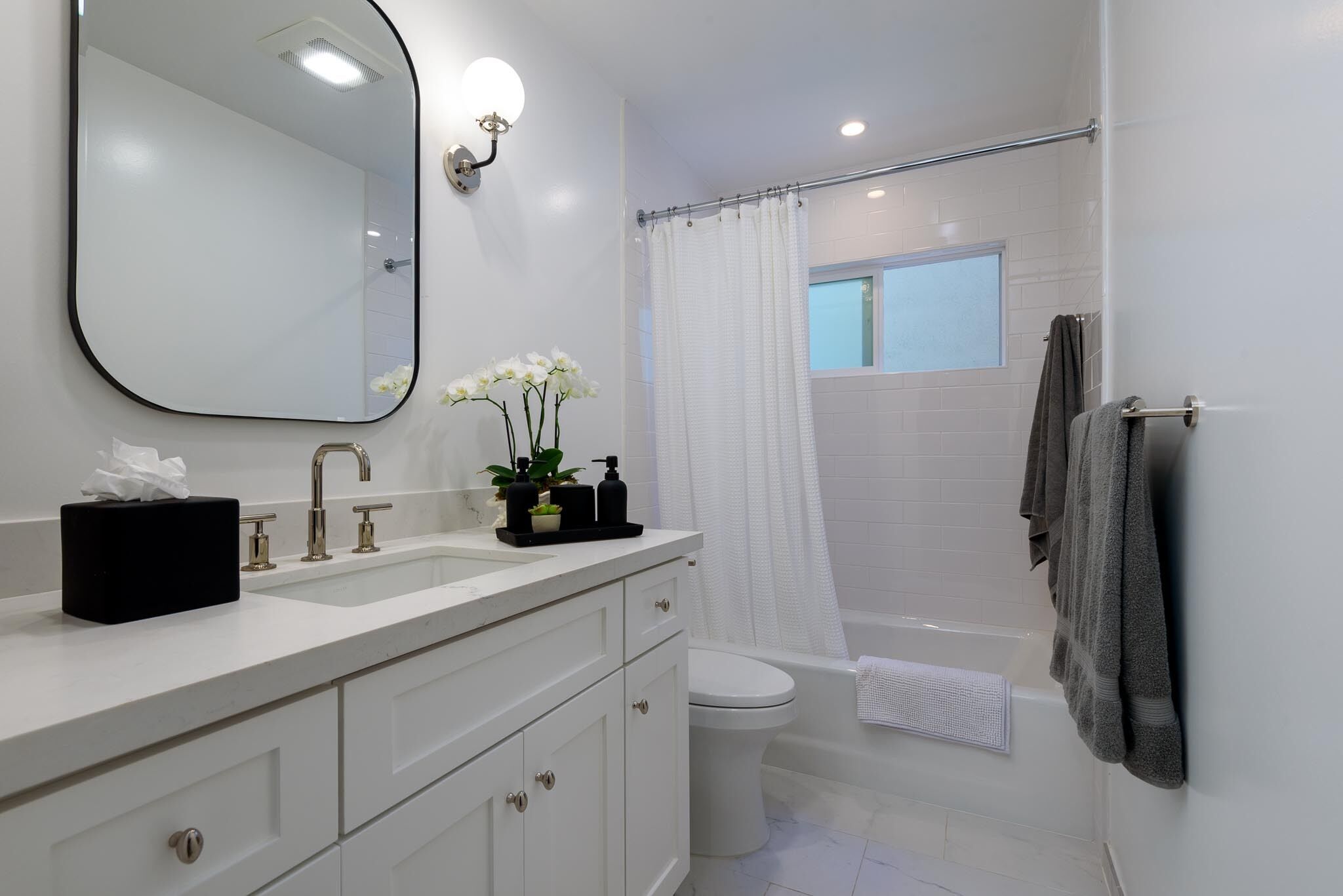 Een moderne badkamer met een witte wastafel, spiegel en toilet. Er is een douchegordijn, een handdoekhouder en een klein raam boven het toilet.