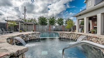 Une piscine aux murs en pierre et agrémentée d’une cascade, entourée d’une clôture en bois et bordée d’un bâtiment en briques.