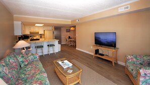 A living room with a sofa, coffee table, TV, and a kitchen area in the background. - Relax in this 2 BD/2Bath Direct Ocean Front. Listen to the ocean waves and enjoy. (OCEAN CITY)