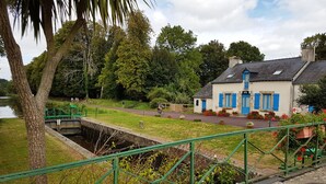 Una casa con contraventanas azules, un pequeño jardín con plantas en macetas y un camino que lleva a un puente sobre un canal de agua.