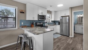 A modern kitchen with white cabinets, a stainless steel refrigerator, a microwave, and a stove. There are two bar stools and a window with blinds. - Beachy, Modern & Updated - Seagrove Beach on 30A + Free Attraction Tickets! (Santa Rosa Beach)