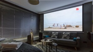 Une chambre d’hôtel moderne avec une grande fenêtre donnant sur une plage, un canapé gris, un lit recouvert d’une courtepointe noire et une petite table basse.