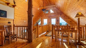 L’intérieur d’un chalet en bois, avec un plafond haut, un plancher en bois, un coin repas et une vue sur les montagnes.