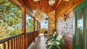 A wooden porch with a view of trees and mountains, a stone wall, and a mounted animal head. - Luxury Log Cabin ❃ Panoramic View ❃ Sauna ❃ Starlink ❃ 15 miles to National Park (Luray)
