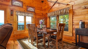 L’intérieur d’un chalet en bois présente une table à manger dressée pour quatre personnes, entourée de chaises en bois. On y trouve deux fenêtres ornées de rideaux à carreaux, une horloge et des tableaux encadrés accrochés aux murs.