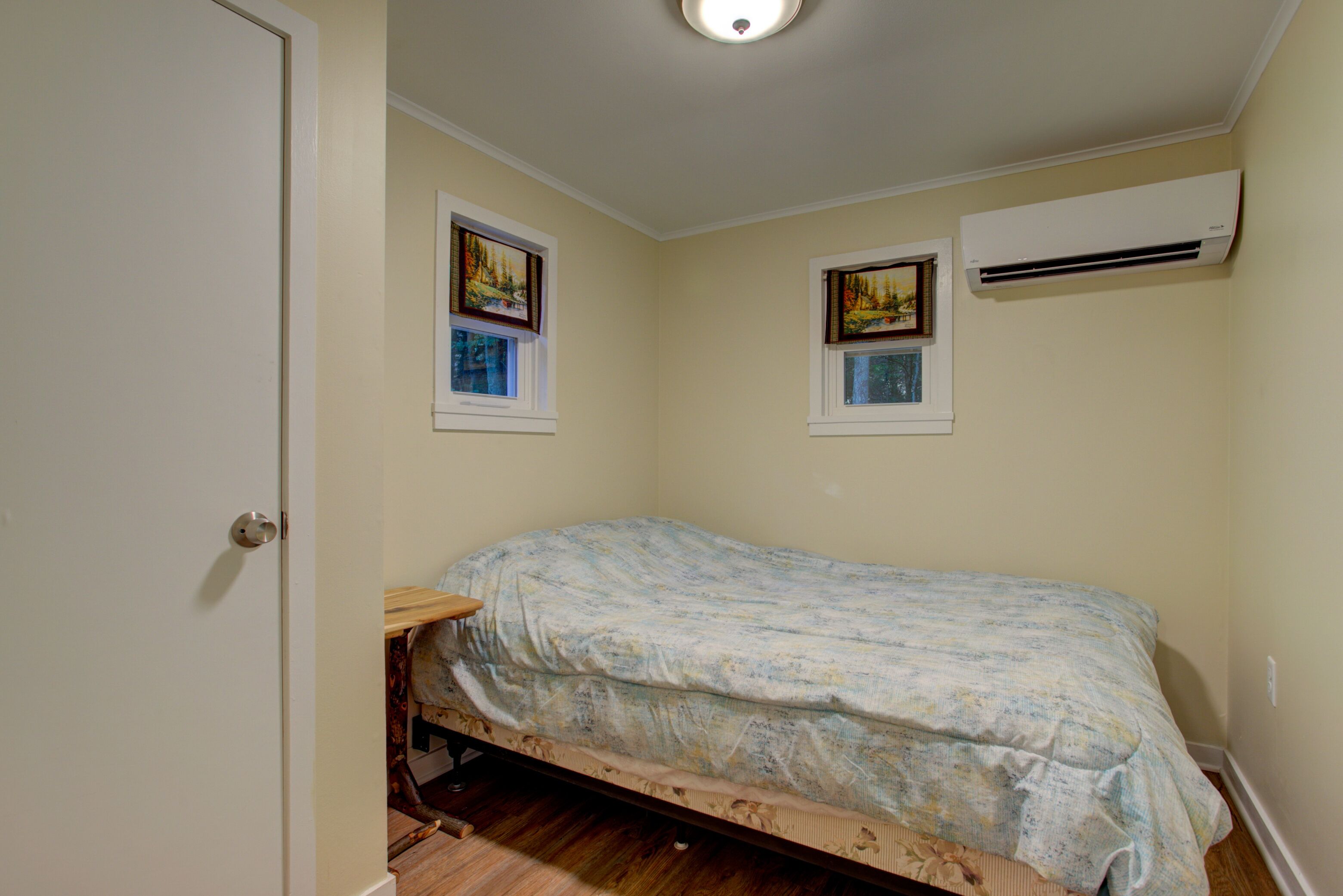 A small bedroom with a single bed, two windows, and an air conditioning unit.