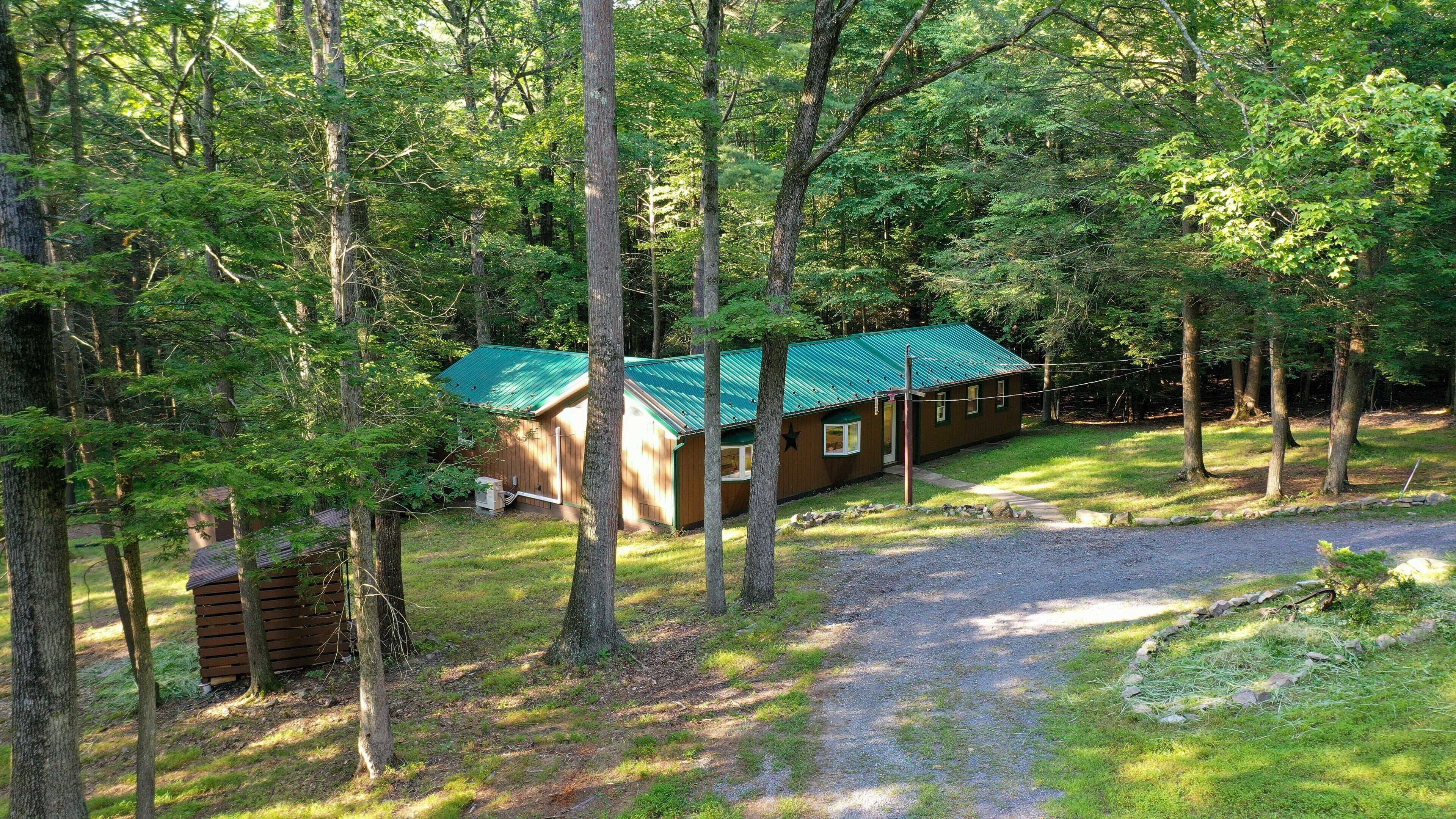 A cabin with a green roof surrounded by trees, a gravel path, and a small grassy area.