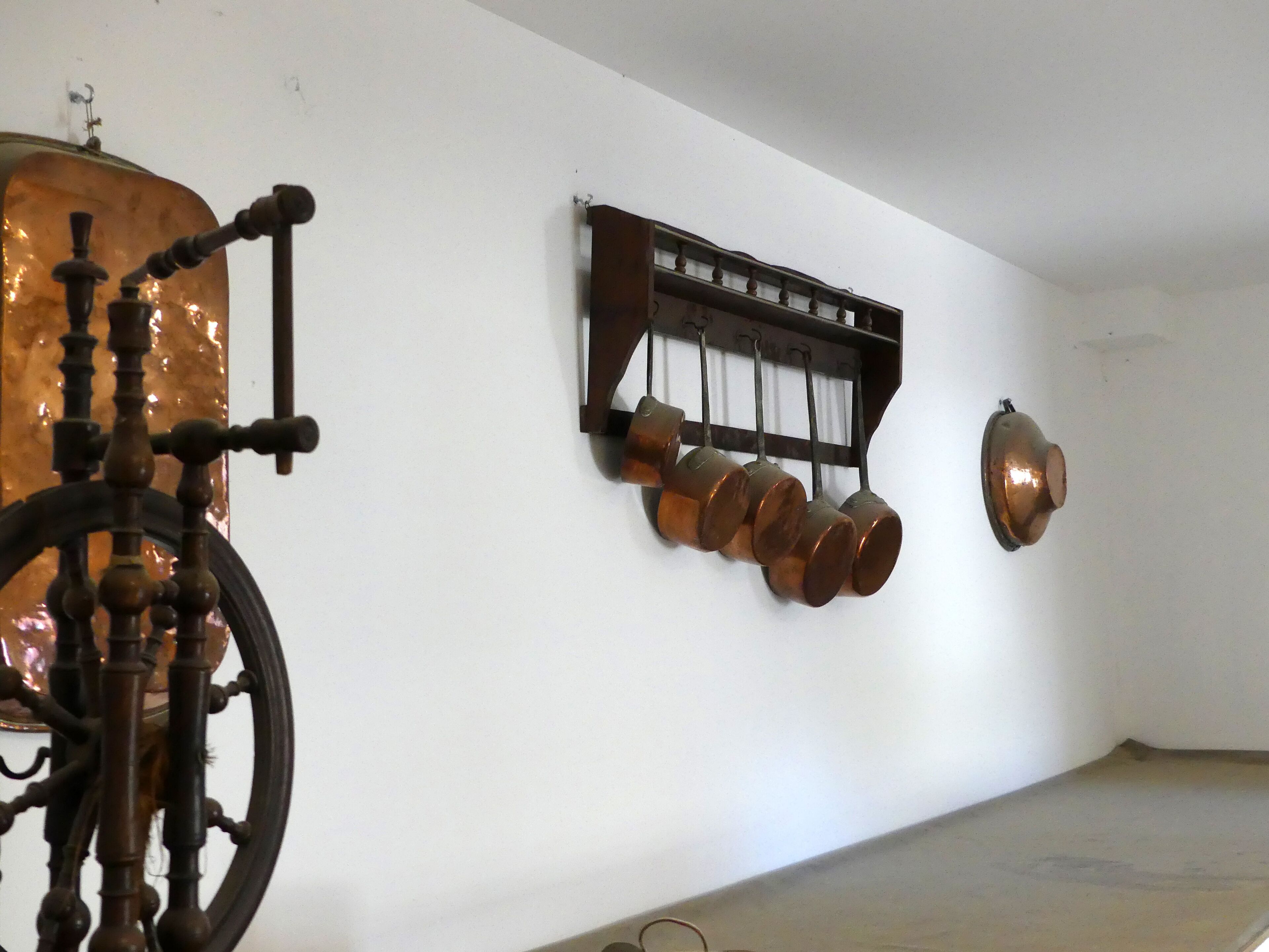 A room with a large copper still, a wooden barrel, and a wall-mounted rack with copper pots.